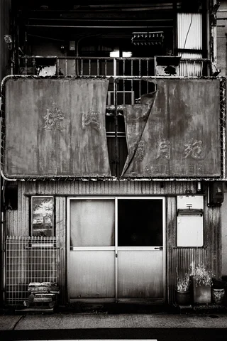 Torn balcony coverings hang above sliding doors on a weathered narrow facade in a black and white view.