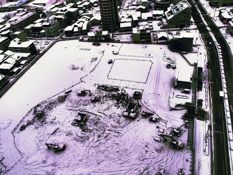 Snow blankets a broad construction site and surrounding rooftops beside a curving canal in black and white.