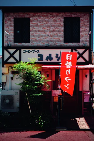 A red vertical banner stands outside a narrow cafe with brick-patterned upper walls and a red entrance.