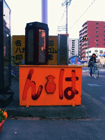 A bright orange sign with rounded red Japanese characters stands beside a utility pole as a cyclist approaches.