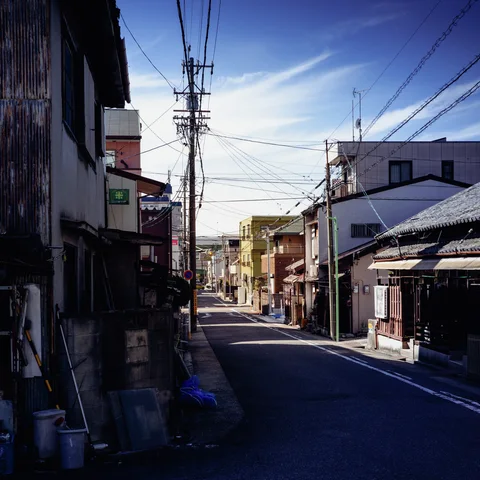 A quiet narrow street runs between low buildings beneath utility poles and crossing wires.