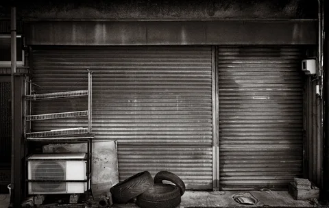 Discarded tires and metal panels rest before closed corrugated shutters in black and white.