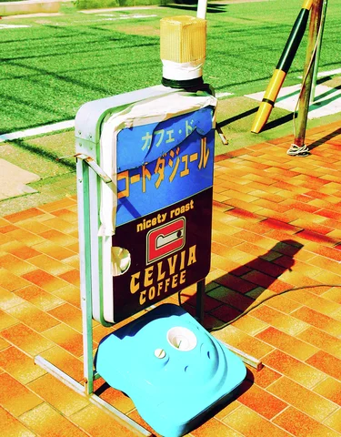 A freestanding blue cafe sign on a turquoise weighted base casts a long shadow across orange tiles.