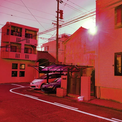 A curving residential street passes parked cars and a covered driveway amid red-toned buildings and overhead wires.