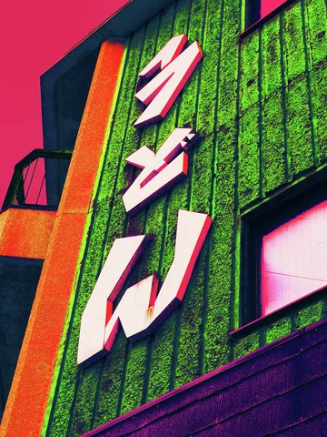 Large pale Japanese characters project from a green vertically textured facade beside orange and pink trim.