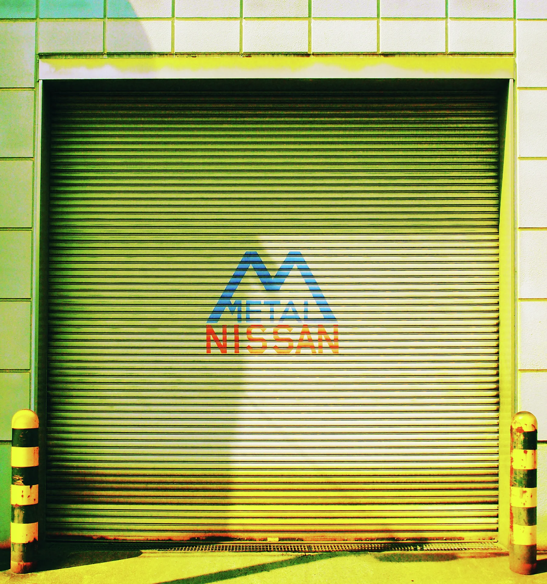 A blue angular emblem marks a yellow-green closed shutter between striped protective bollards.