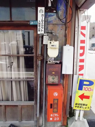 Signs, utility meters and an orange vertical box crowd a weathered wooden storefront beside a sliding window.