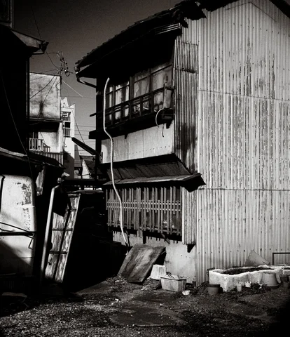 A weathered corrugated house stands beside a narrow cluttered gap with leaning ladders in black and white.