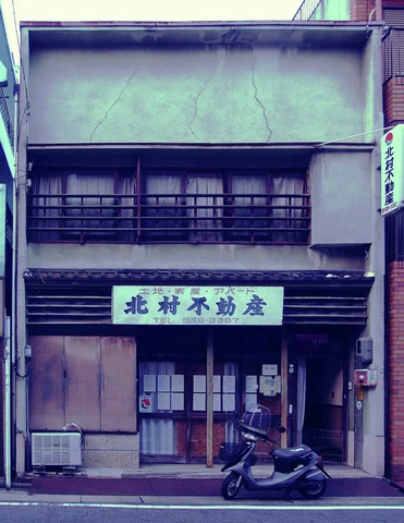 A scooter stands outside a weathered narrow shop beneath a pale sign and barred upper windows.