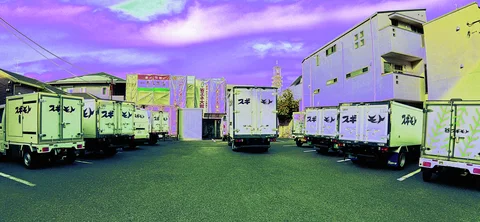 Rows of white delivery trucks fill a paved lot between low buildings beneath purple clouds.