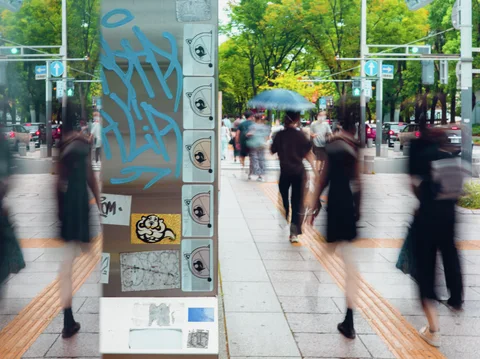 Blurred pedestrians flank a sticker-covered column with blue graffiti beside mirrored glass along a leafy sidewalk.