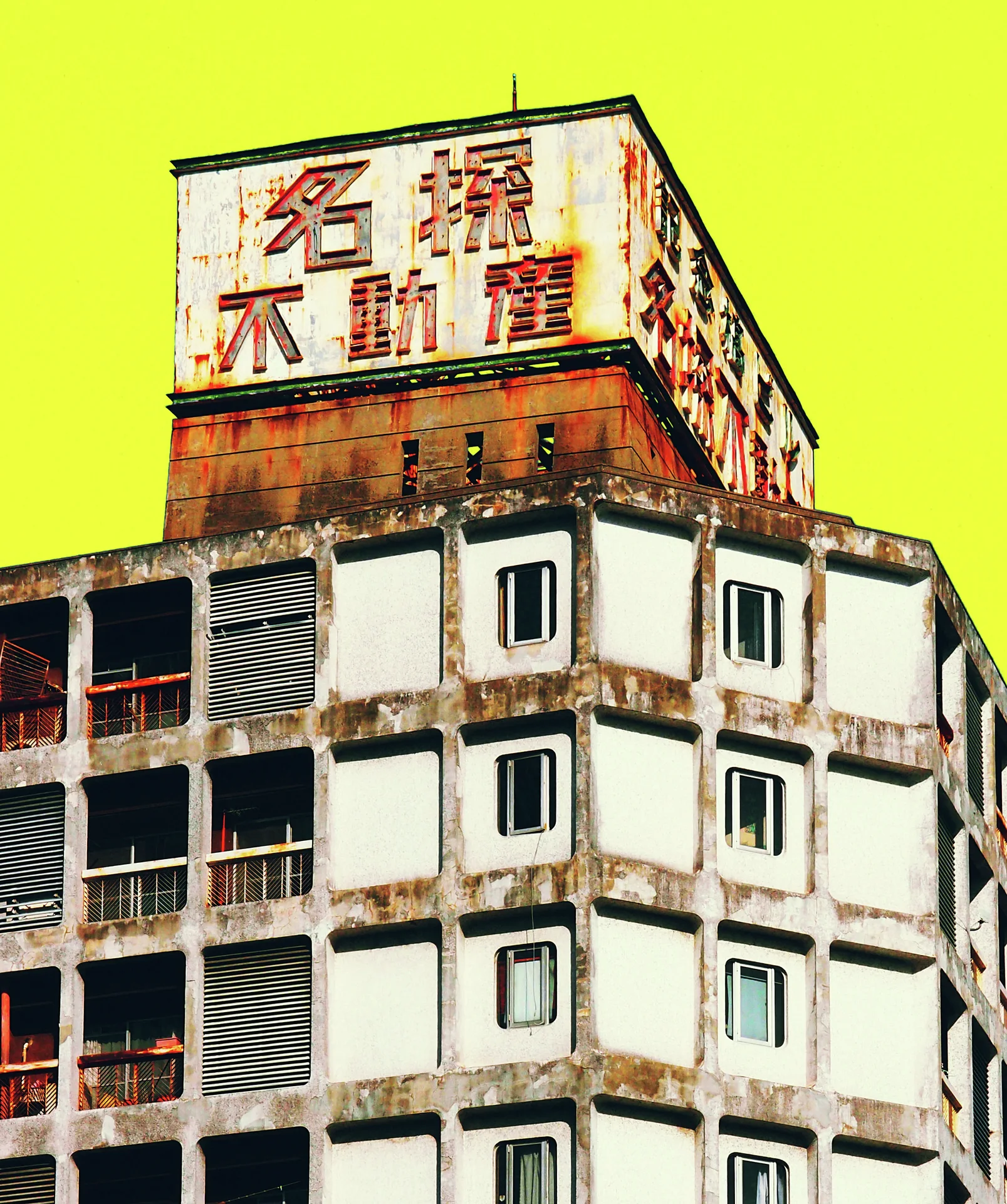A weathered multi-story building with small square windows carries a large rooftop sign against yellow sky.