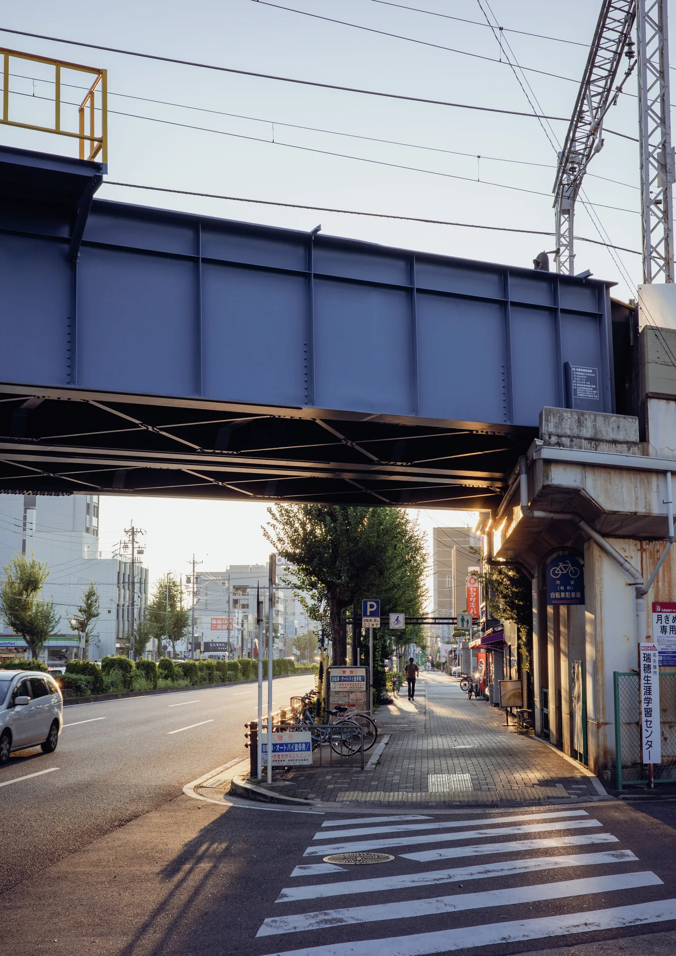 A blue steel railway bridge spans a road and sunlit sidewalk beside shuttered storefronts.