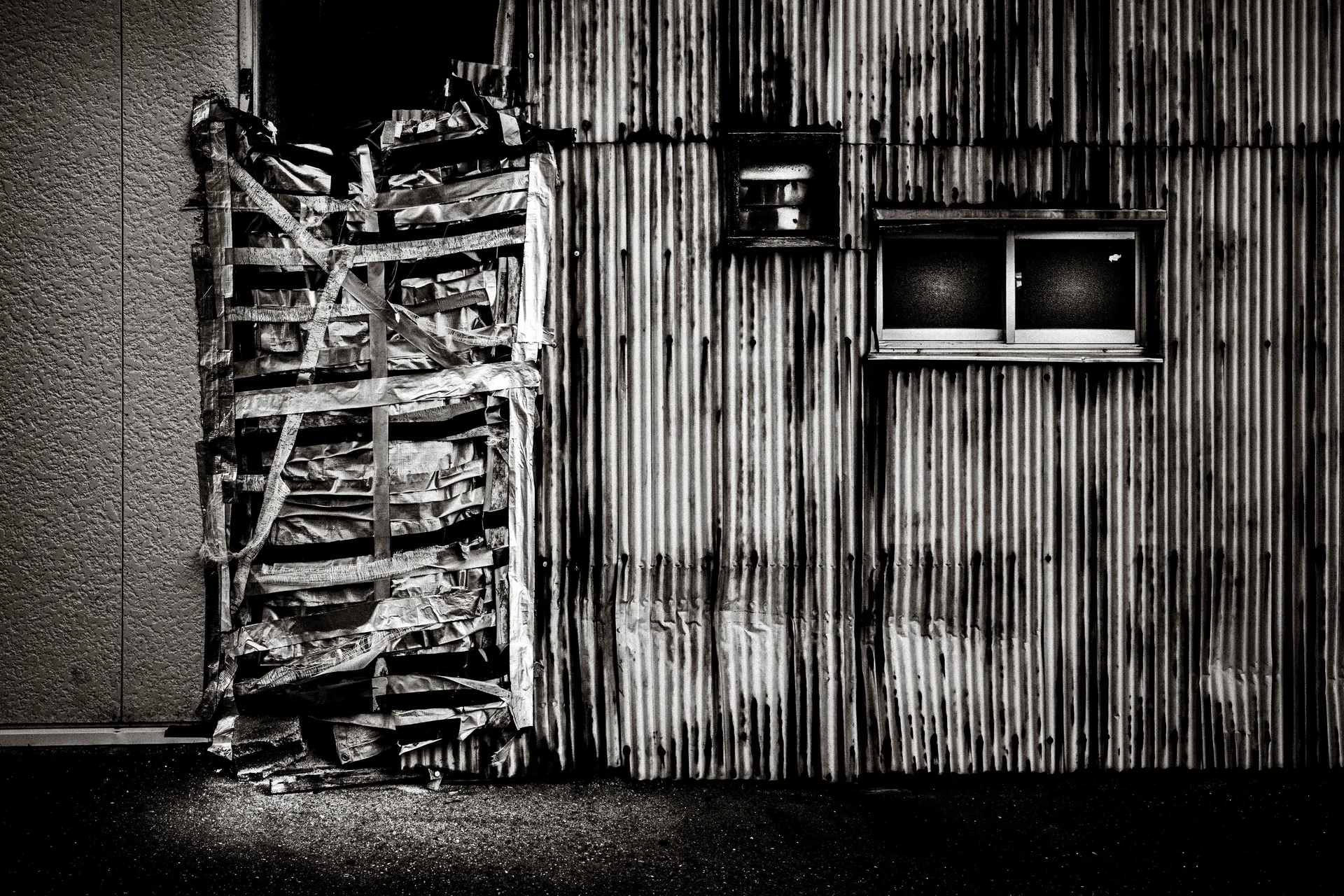 A taped bundle of stacked materials leans beside a corrugated wall with small windows in black and white.