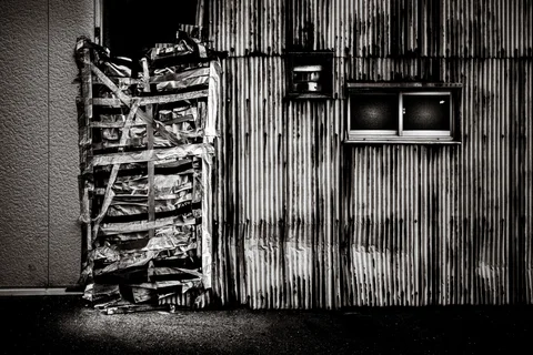 A taped bundle of stacked materials leans beside a corrugated wall with small windows in black and white.