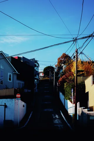 A narrow uphill lane runs between shadowed houses beneath crisscrossing wires and bright blue sky.
