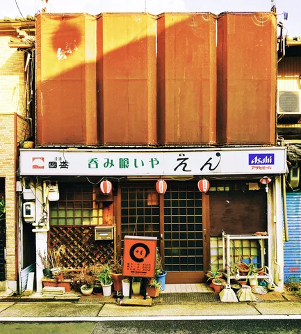 A rust-brown upper facade shelters a small shop with orange lanterns, potted plants, and a white sign.