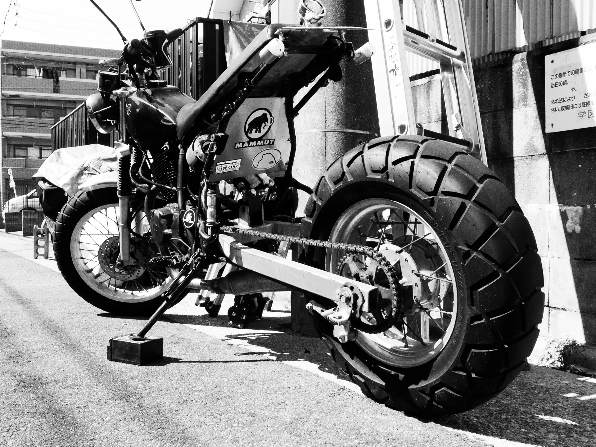 A parked motorcycle with a thick treaded rear tire leans beside a building in bright sunlight.