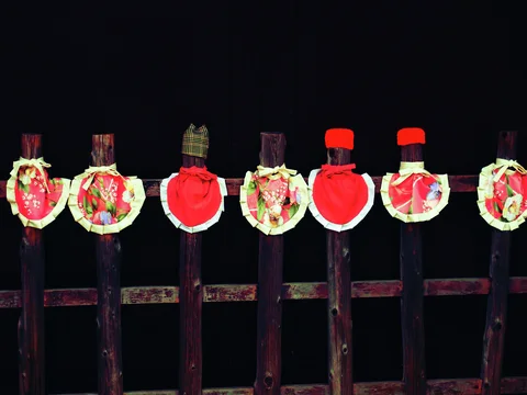 Seven slender wooden figures wear patterned bibs and red cloths against a dark background.