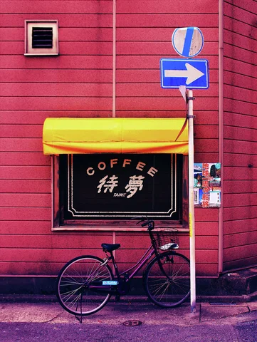 A bicycle leans below a coffee shop window with a yellow awning beside a blue right-arrow sign.