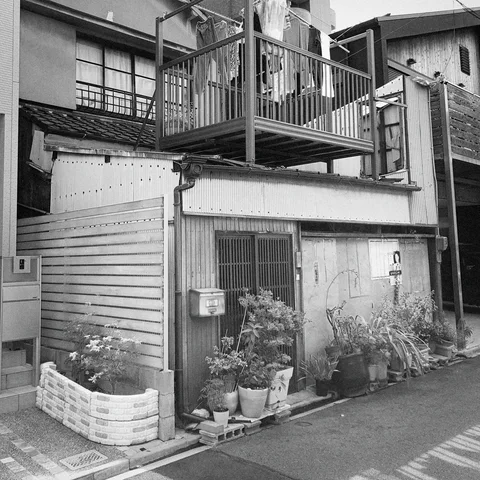 Potted plants line a small weathered residence beneath a raised metal balcony in black and white.
