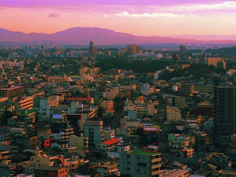 Dense city rooftops extend toward layered distant hills beneath a pink and orange sky.