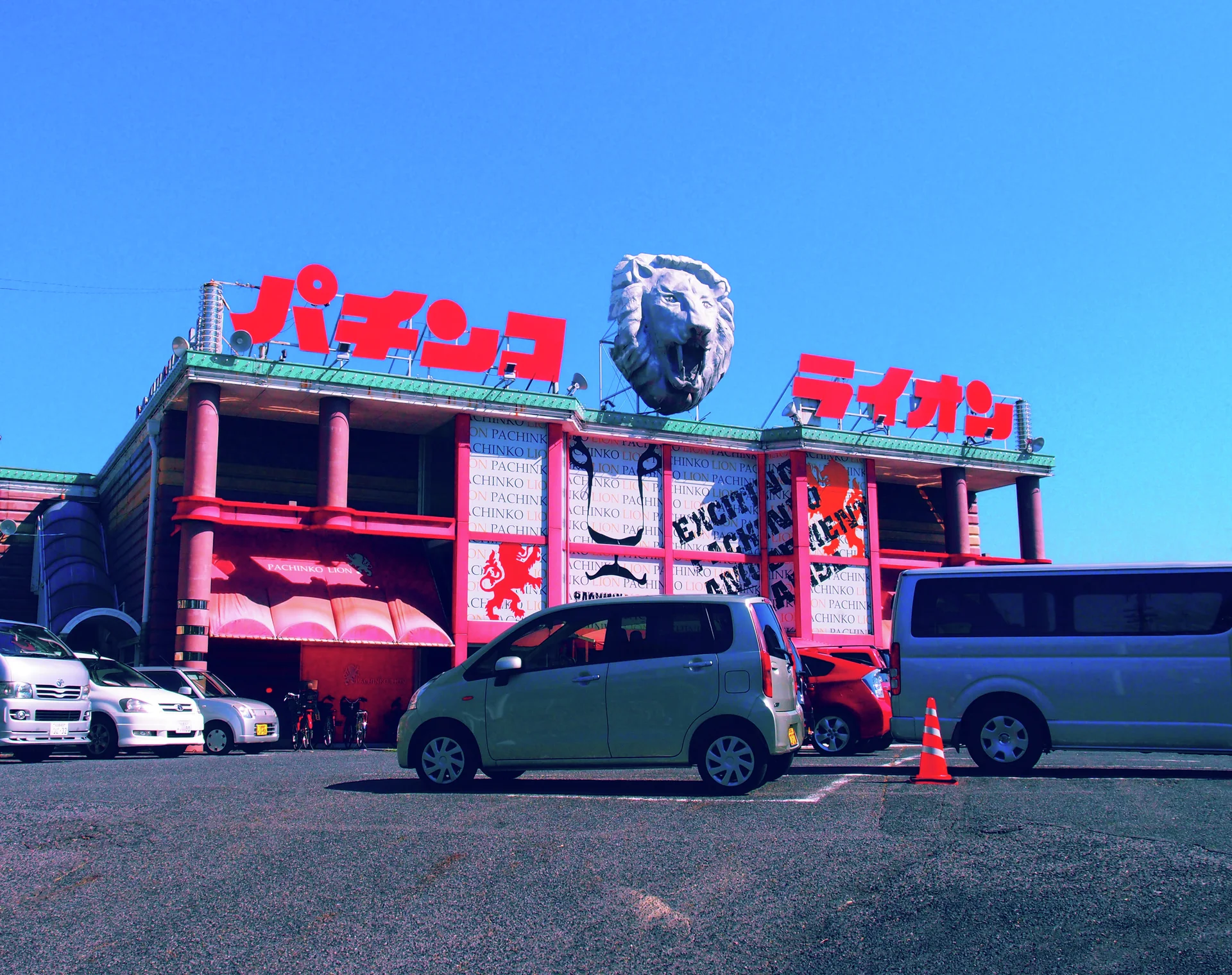 A giant lion head tops a pink commercial building with red Japanese lettering above parked cars and vans.
