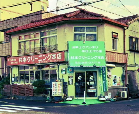 A green corner cleaning shop has large Japanese signs beneath a red tiled roof and overhead wires.