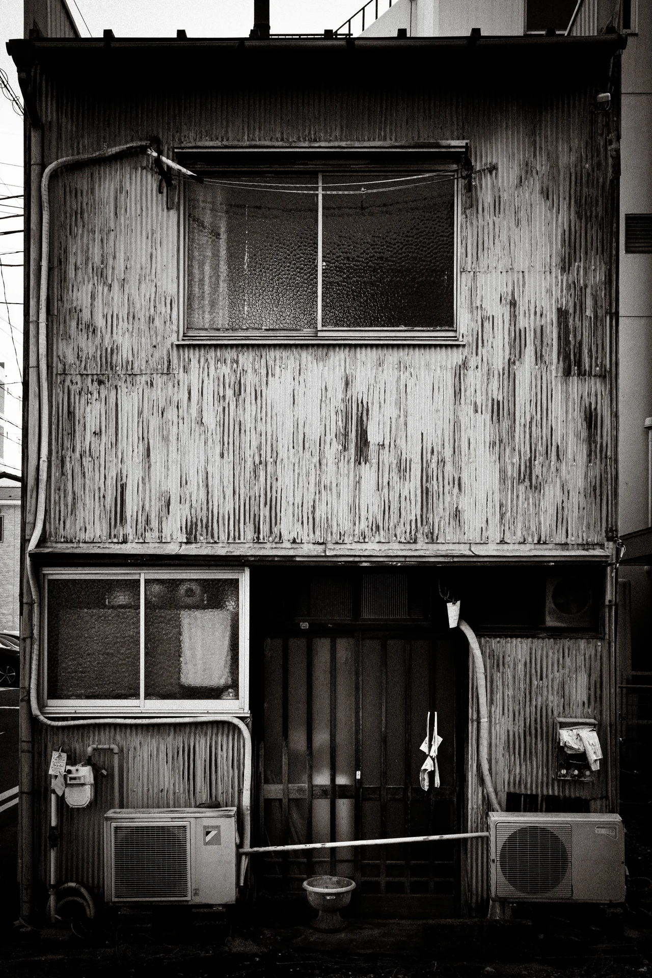 A weathered two-story wooden facade has screened windows and two ground-level air conditioners in black and white.