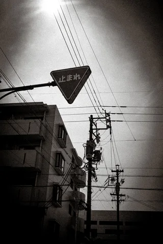 A triangular street sign hangs beneath bright sun and crossing utility wires in black and white.