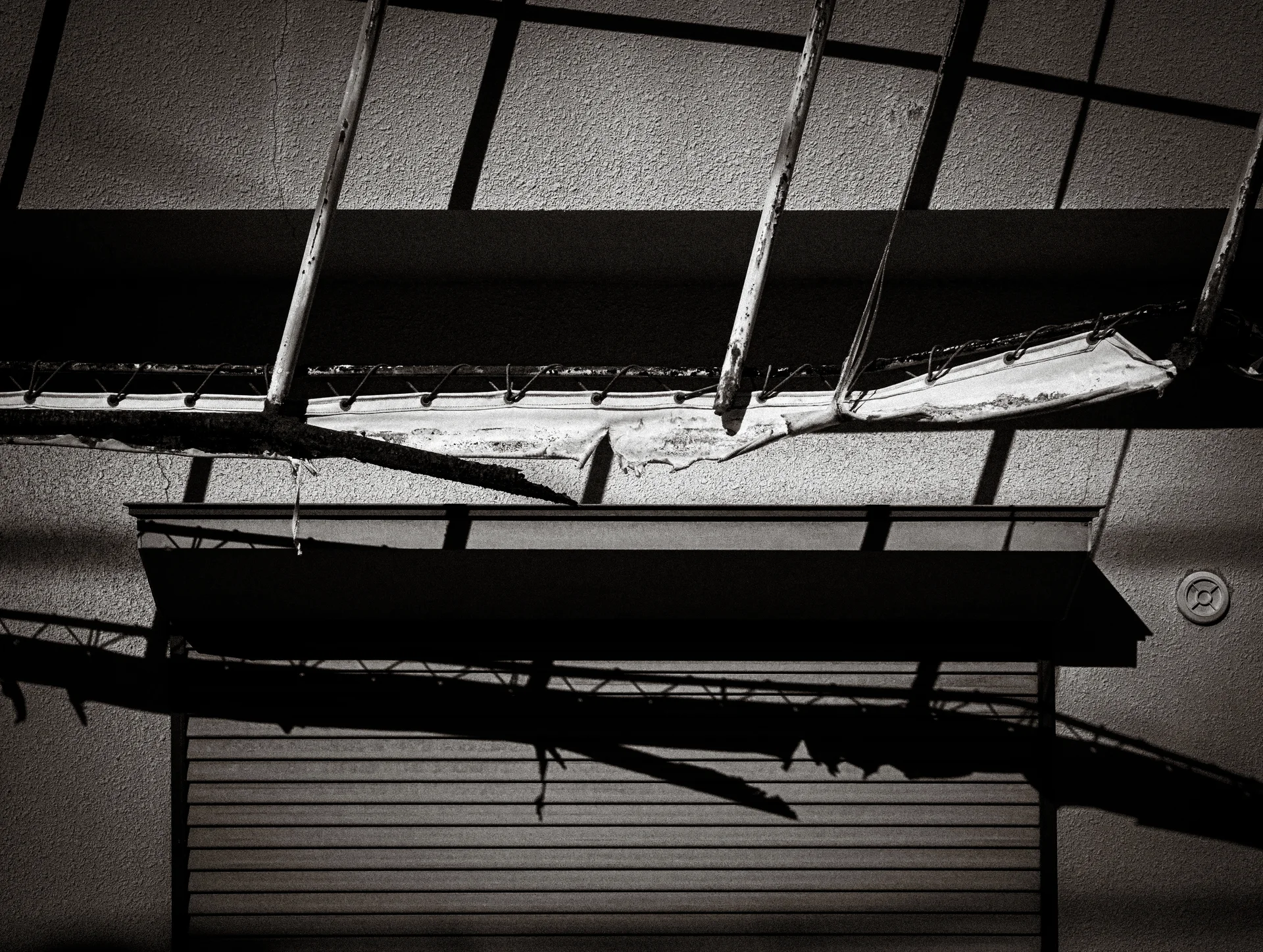 Broken canopy panels hang above a closed shutter with angular shadows in a black and white view.