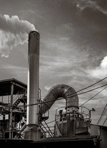 A tall industrial chimney and curved duct rise above metal platforms beneath a cloudy black and white sky.