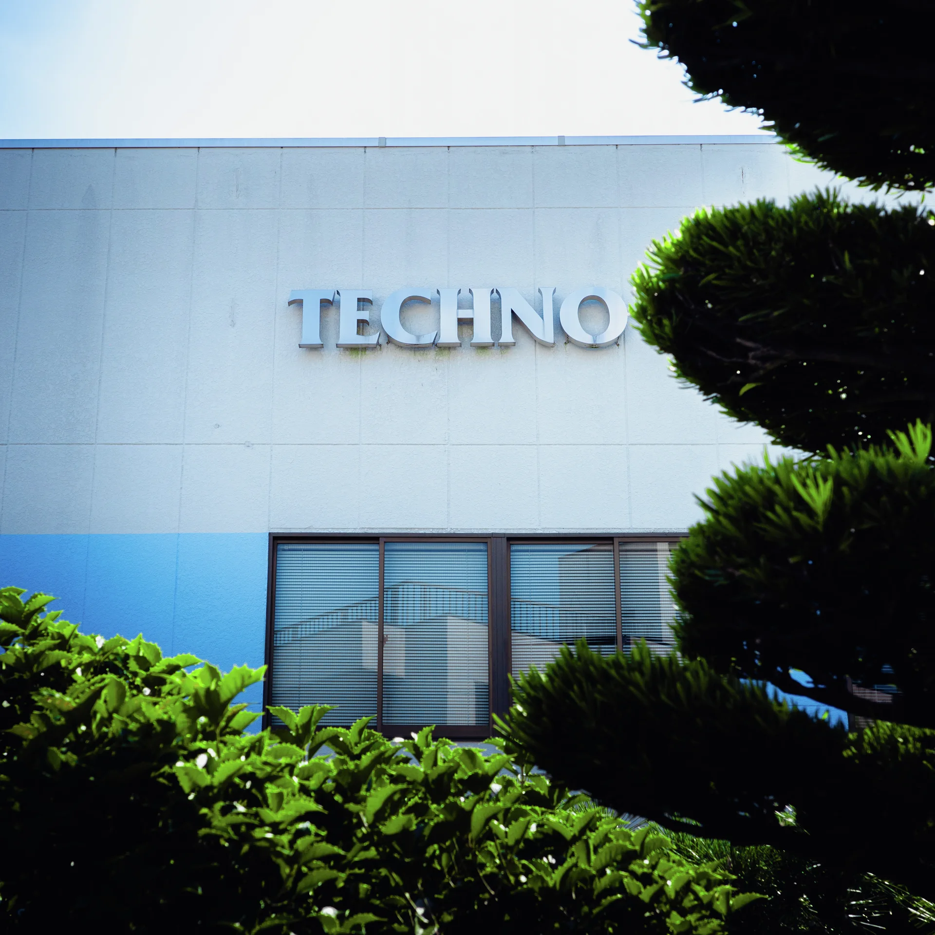 Raised silver lettering marks a pale blue building facade partly obscured by neatly trimmed trees and shrubs.