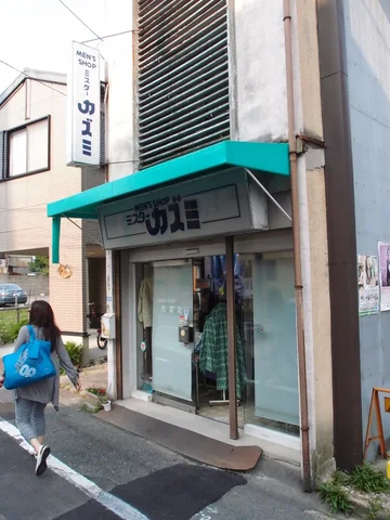 A pedestrian carrying a bright blue bag passes a narrow shop beneath a turquoise awning.