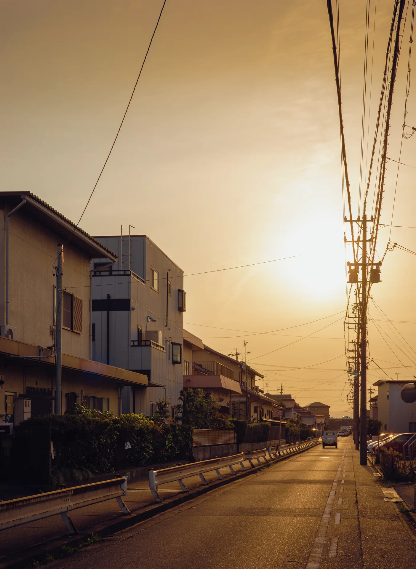 Low sunlight illuminates a quiet narrow street beneath an orange sky crossed by dense overhead wires.