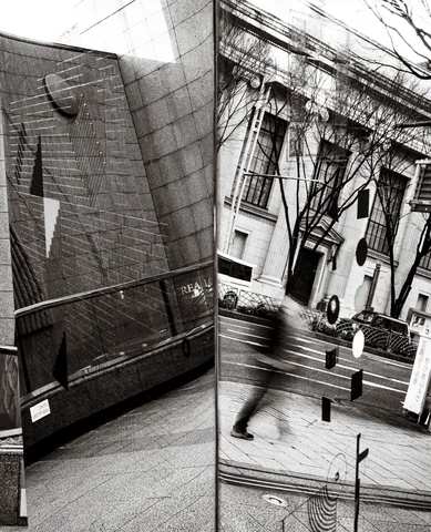 A reflective panel juxtaposes buildings, bare branches and a blurred passerby in black and white.