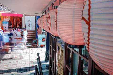 A row of large white paper lanterns hangs beside a covered shopping walkway with blurred pedestrians.