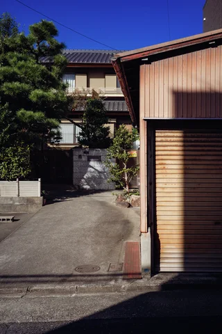 A narrow driveway leads between a shuttered brown building and trees toward a two-story house.