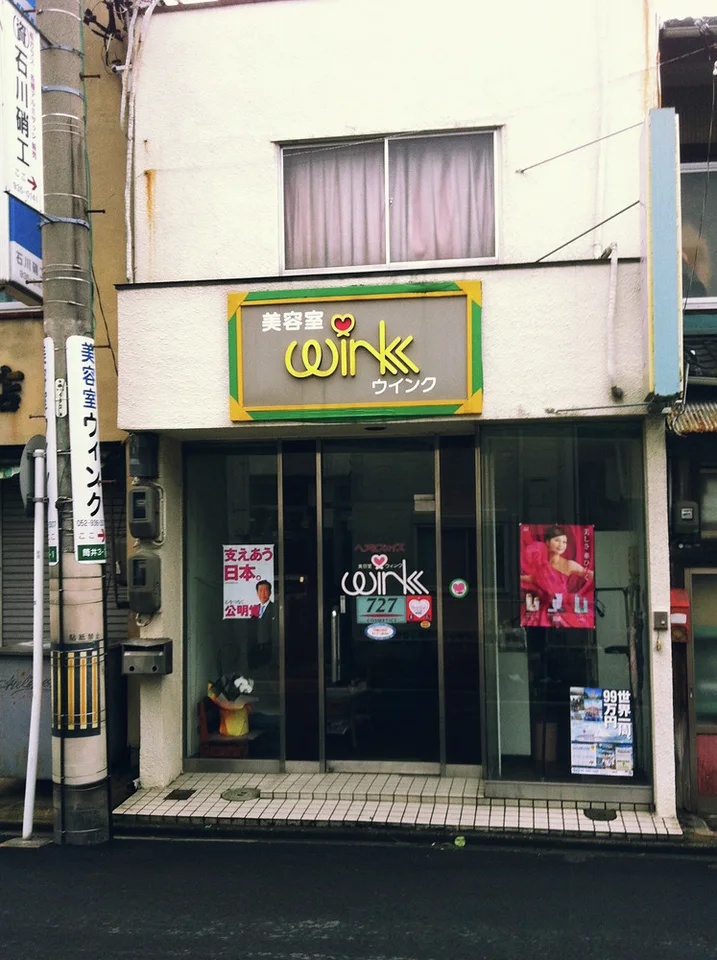 A narrow cream storefront has glass doors beneath a green sign and an upper curtained window.
