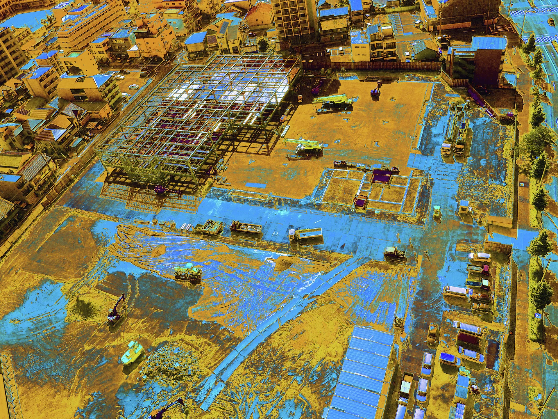 An overhead view shows a broad construction site with steel framing, vehicles, and vivid blue ground coverings.