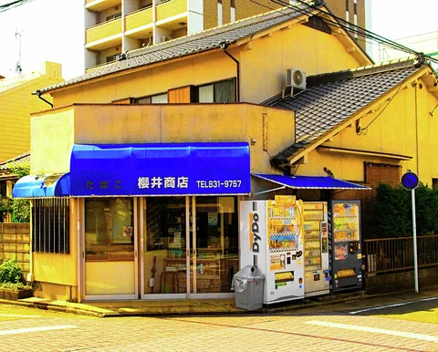 A blue awning shades a yellow corner shop beside drinks vending machines beneath tiled roofs.