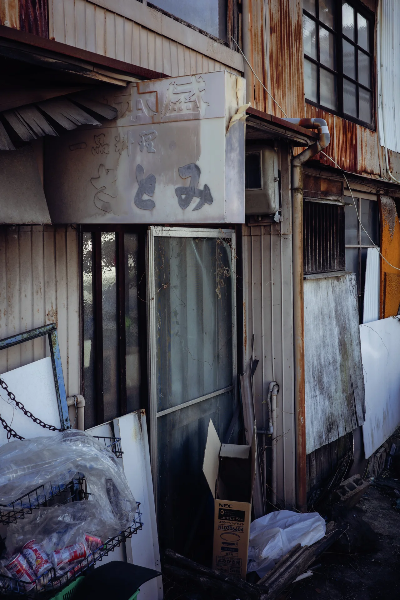 A faded projecting sign hangs above sliding glass doors in a narrow rust-streaked metal facade.