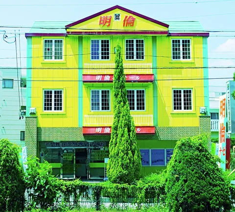 A bright yellow building with green trim and repeated grid windows rises behind neatly shaped shrubs.