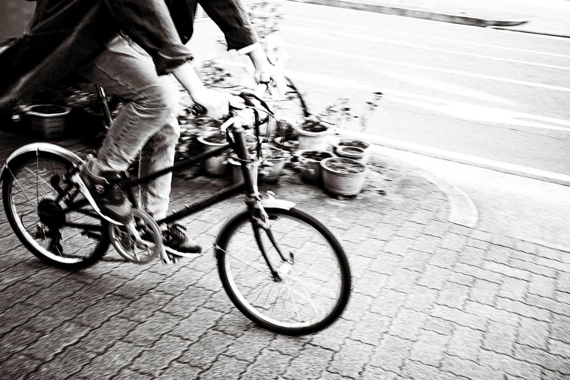 A cropped cyclist rides a small-wheeled bicycle past potted plants in a blurred black-and-white street scene.