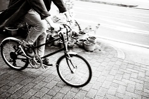 A cropped cyclist rides a small-wheeled bicycle past potted plants in a blurred black-and-white street scene.