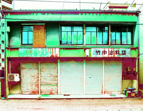 Rust streaks mark a pale green two-story shop above closed metal shutters and dark-framed upper windows.