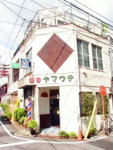 A pale corner barber shop has a red diamond facade panel and striped pole beside its entrance.