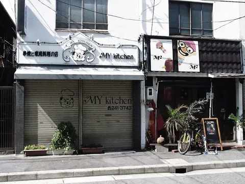 Two small adjacent storefronts have illustrated signs, a closed shutter, and a bicycle beside plants.