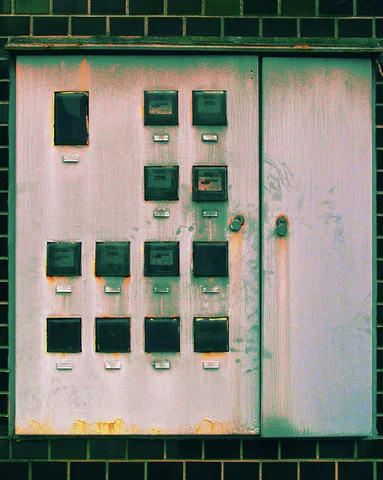Rows of small dark electrical meters dot a weathered pale pink panel within a black tiled wall.