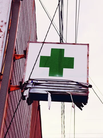 A green cross marks a white projecting sign beneath overhead wires beside a corrugated wall.
